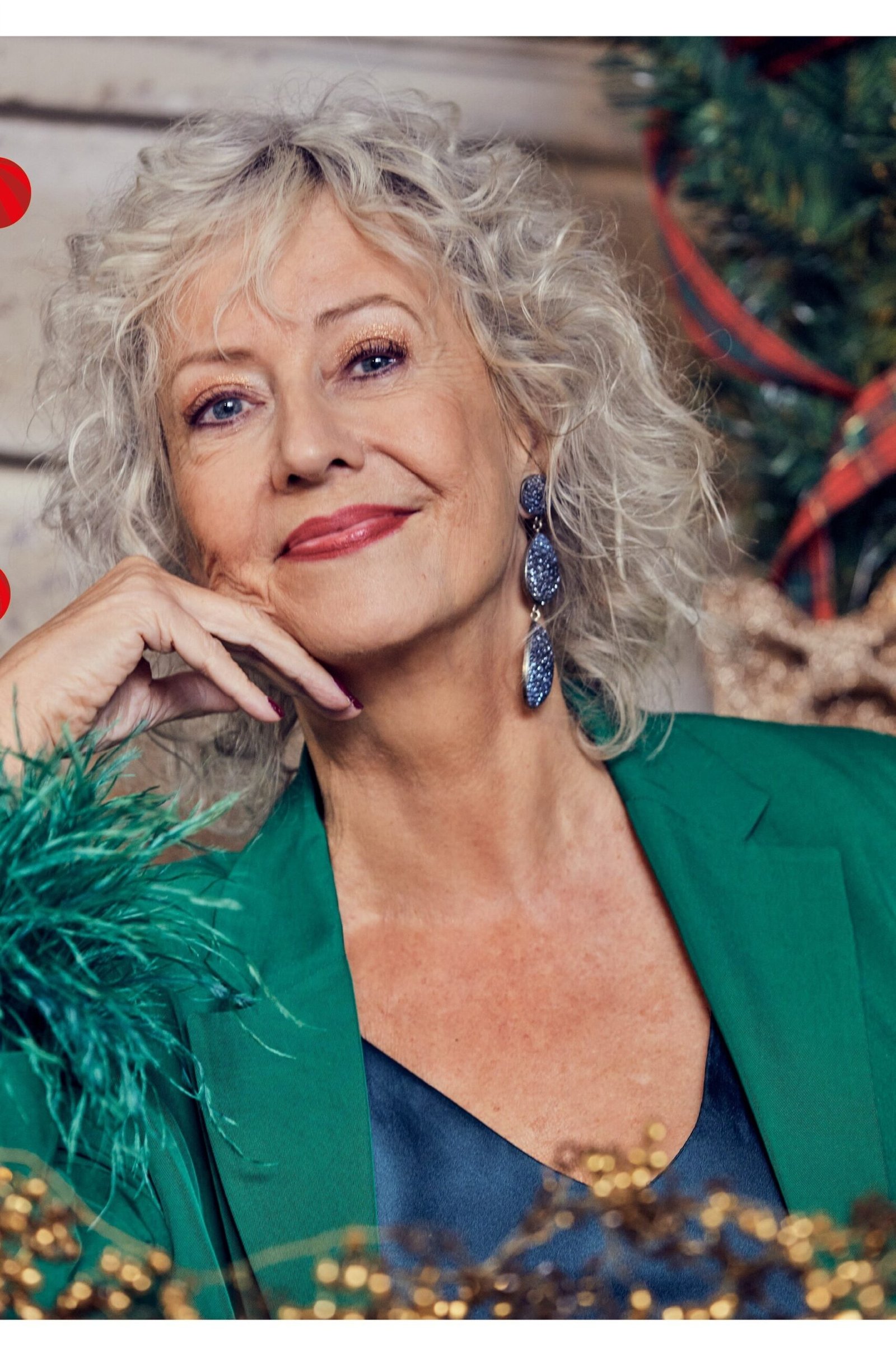 Woman with curly gray hair in emerald-green blazer with blue earrings near Christmas garland, jewel-toned New Year's Eve blazer outfit for women over 60