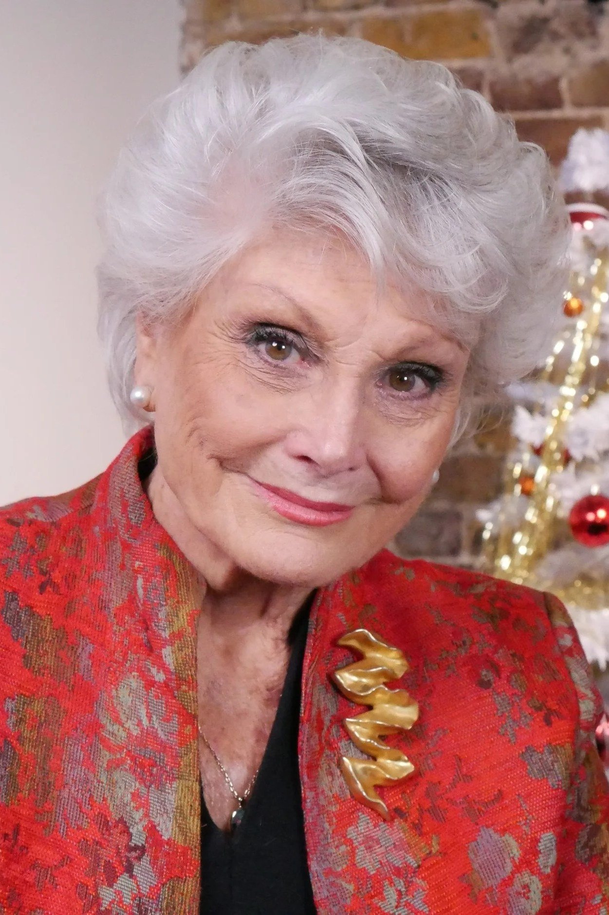 White-haired woman in red floral brocade blazer with gold brooch by Christmas tree, vintage-glam New Year's Eve blazer outfit for women over 60