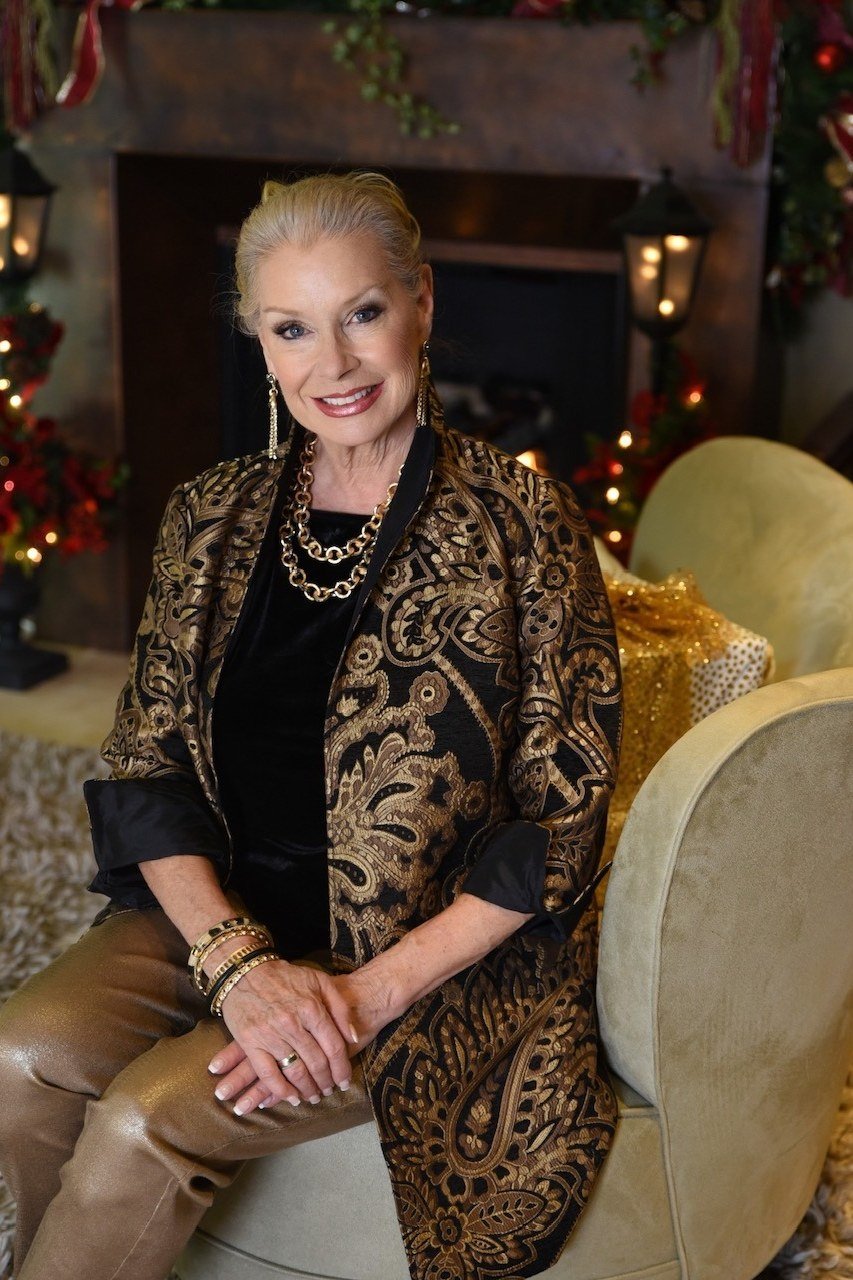 Blonde woman in gold brocade blazer with brown pants seated by fireplace, cozy New Year's Eve blazer outfit for women over 60