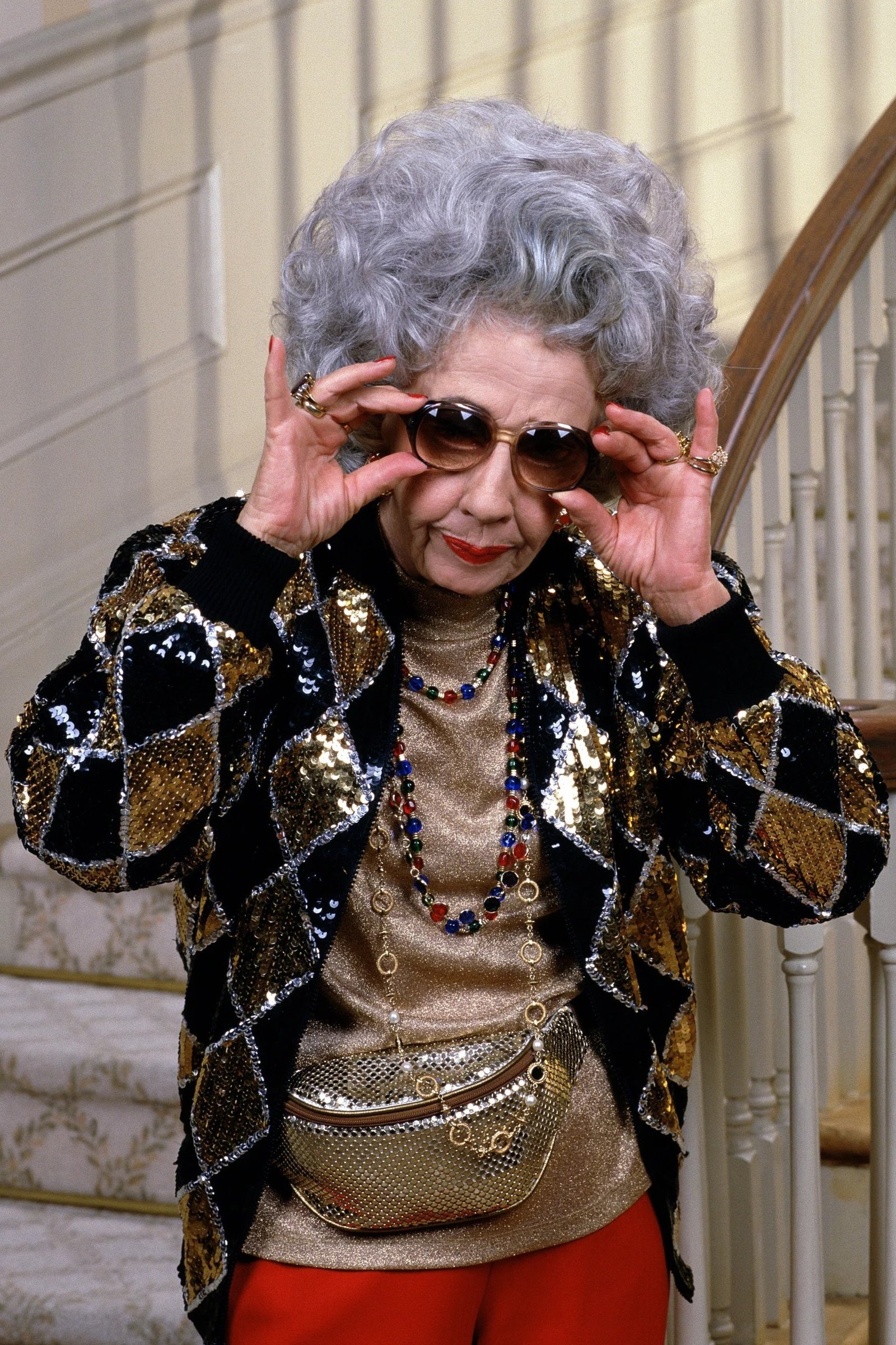 Gray-haired woman in black-and-gold sequin blazer with sunglasses on staircase, art-deco New Year's Eve blazer outfit for women over 60
