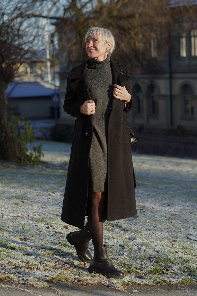 Black long coat over olive green sweater dress with tights and ankle boots, winter layering look for women over 60