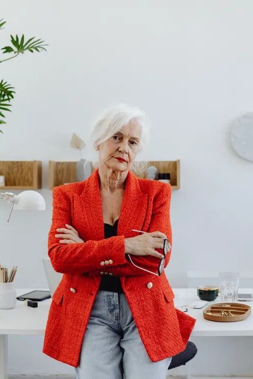 White-haired woman in red blazer, white top and jeans at an office desk
