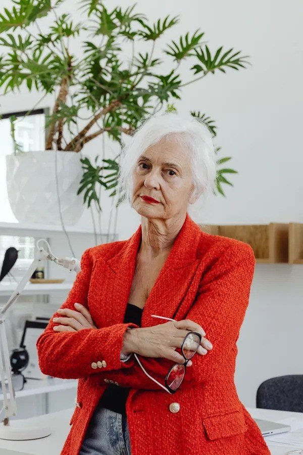 Older woman in red textured blazer, white top and jeans in a bright office