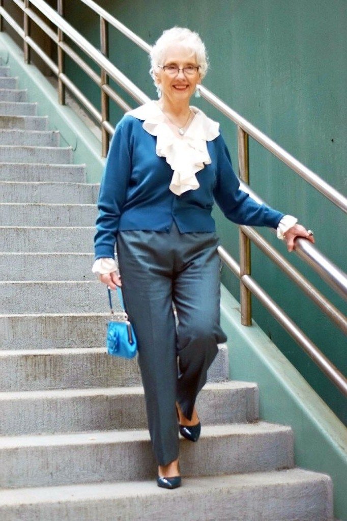 Older woman in blue cardigan, white ruffle blouse and gray trousers on stairs