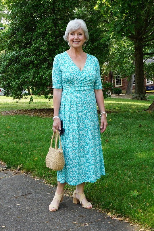 Gray-haired woman over 60 wearing a turquoise floral wrap midi dress with straw bag in a park
