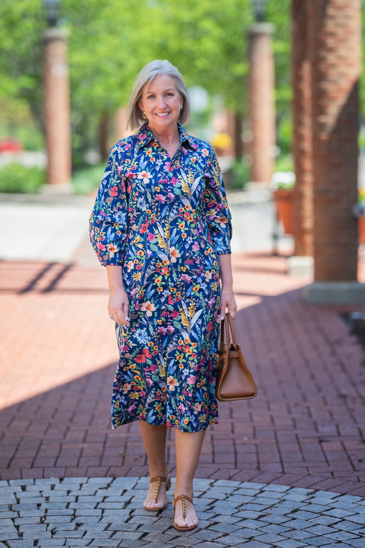 Gray-haired woman over 60 wearing a blue floral wrap midi dress on a brick street