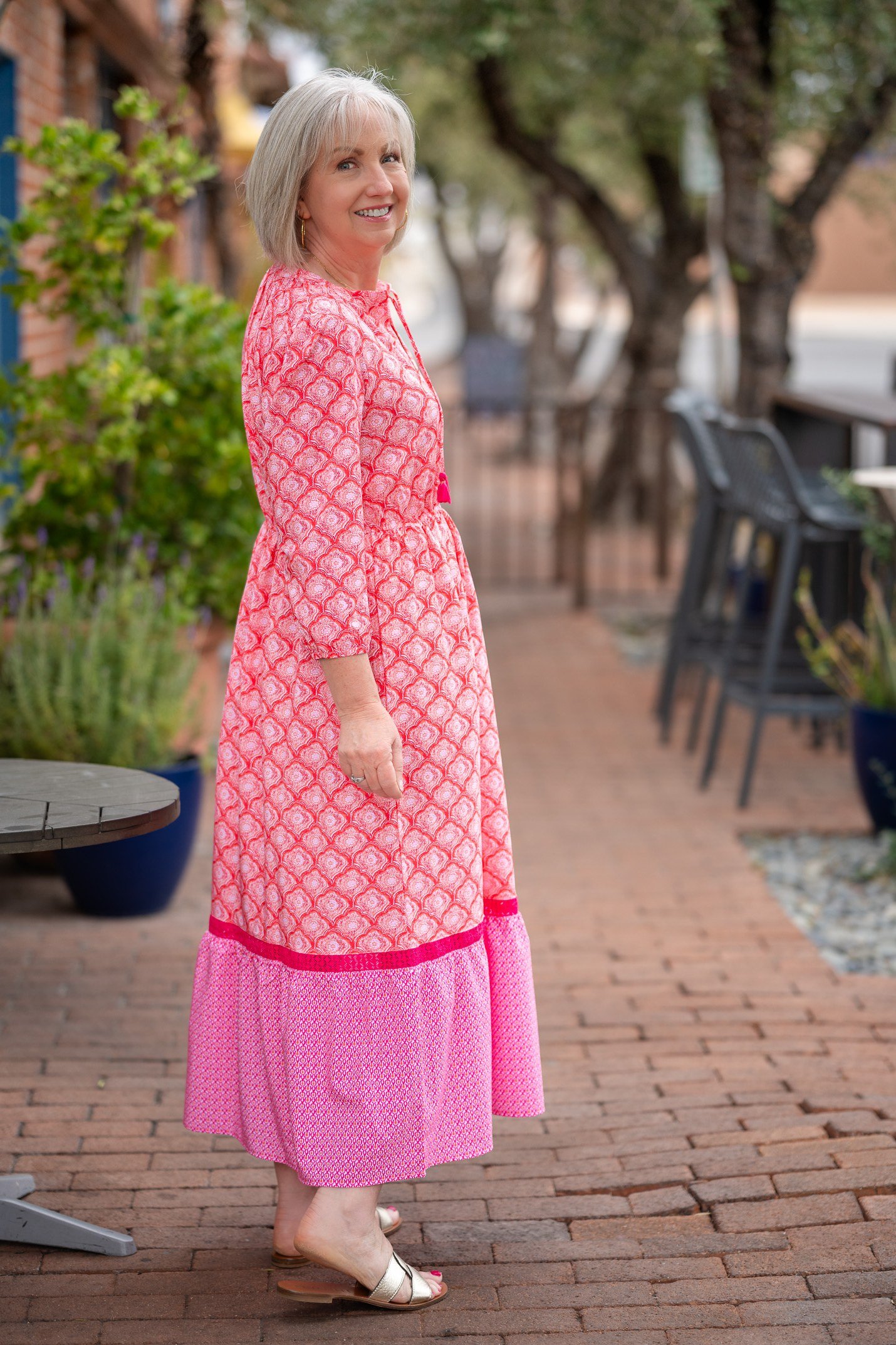 Gray-haired woman over 60 wearing a pink patterned wrap maxi dress on a brick path