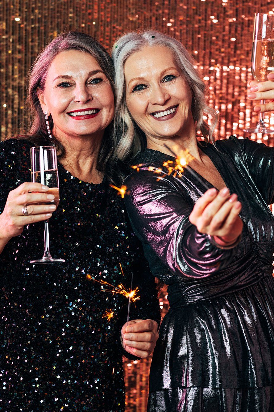 Two older women in gold sequin dresses toasting champagne, Christmas party outfits for women over 70