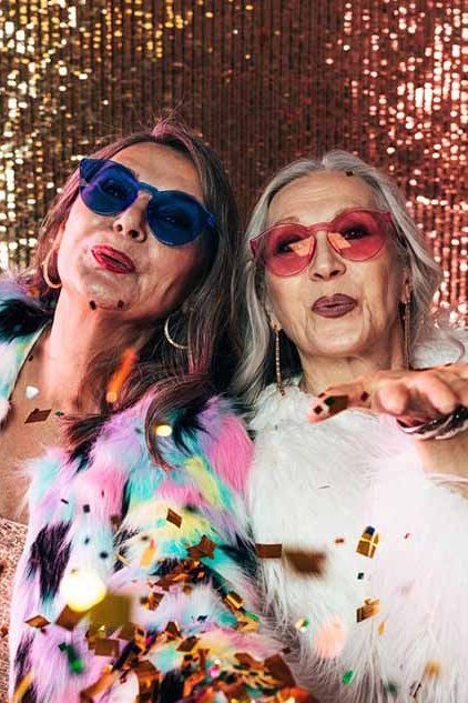 Two elderly women in white and multicolor feather jackets with gold sequin backdrop, glamorous Christmas outfits for women over 70