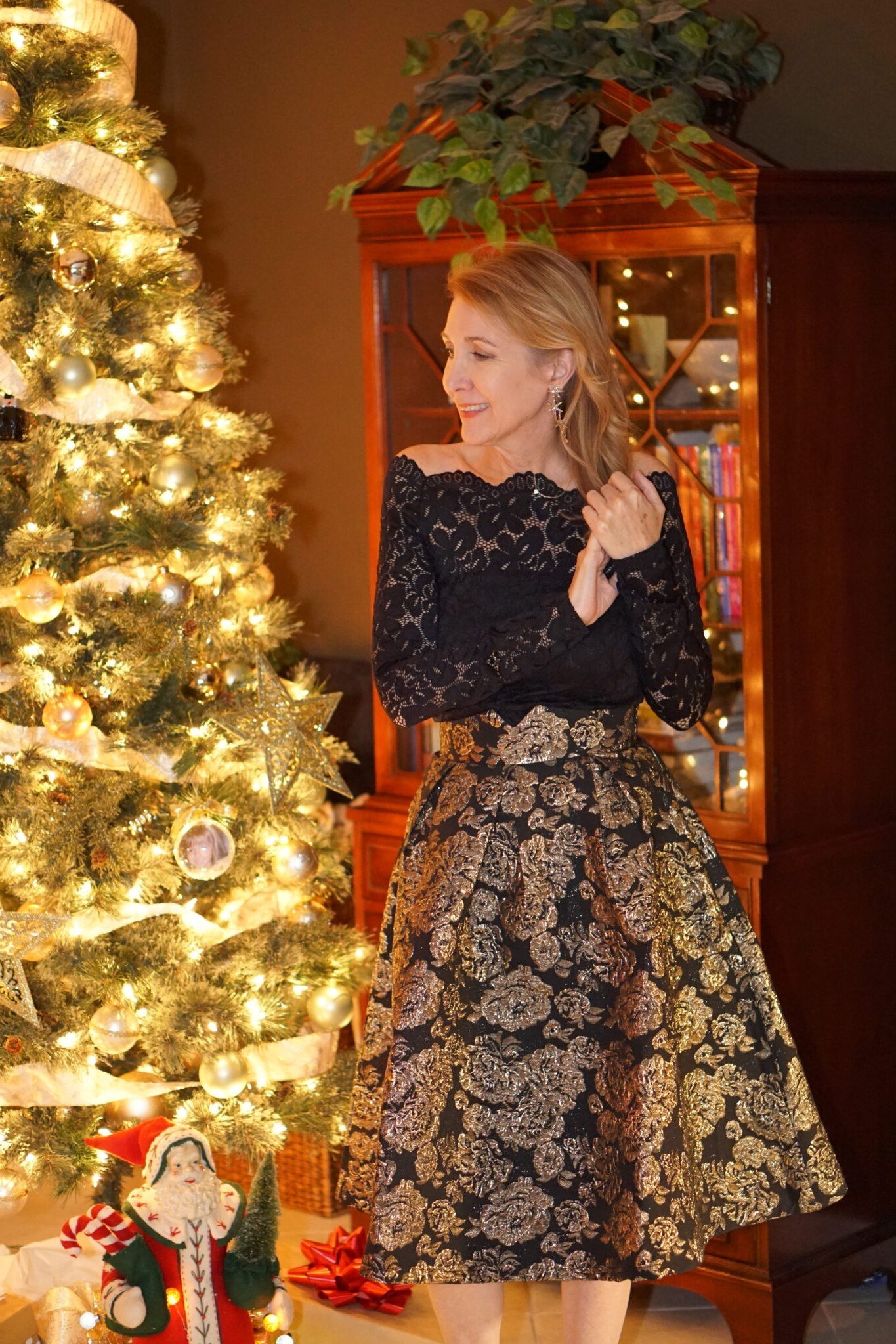 Black off-shoulder lace top with gold and black brocade midi skirt by lit Christmas tree, glamorous holiday outfit for women over 70