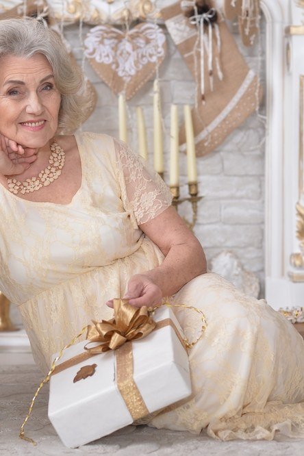 Cream satin Christmas gown reclining with gift box by tree, dreamy holiday outfit for women over 70